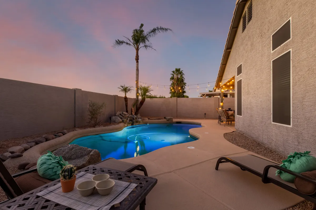 A tranquil backyard scene featuring a sparkling swimming pool, palm tree, comfortable lounge chairs, and lush decorative plants. String lights are draped above, creating a cozy ambiance for evening gatherings or relaxing after a day of adventure.