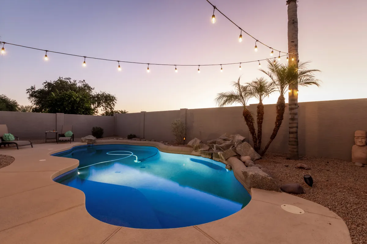 A serene poolside area accented by hanging lights, lush landscaping, palm trees, and a decorative statue. The inviting water and lounge spaces are ideal for both relaxing mornings and lively evening gatherings.
