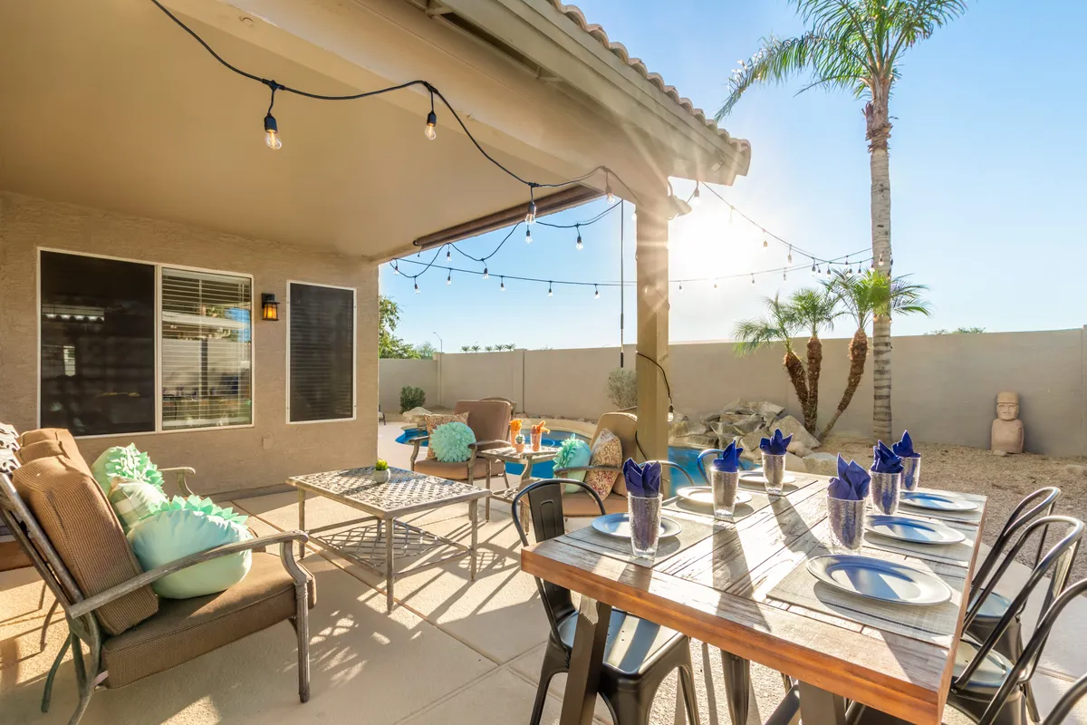 The image features an outdoor dining area set for a meal, complete with decorative pillows, string lights, and palm trees in the background. The space feels effortlessly casual and ready for group dinners or celebratory gatherings at sunset.