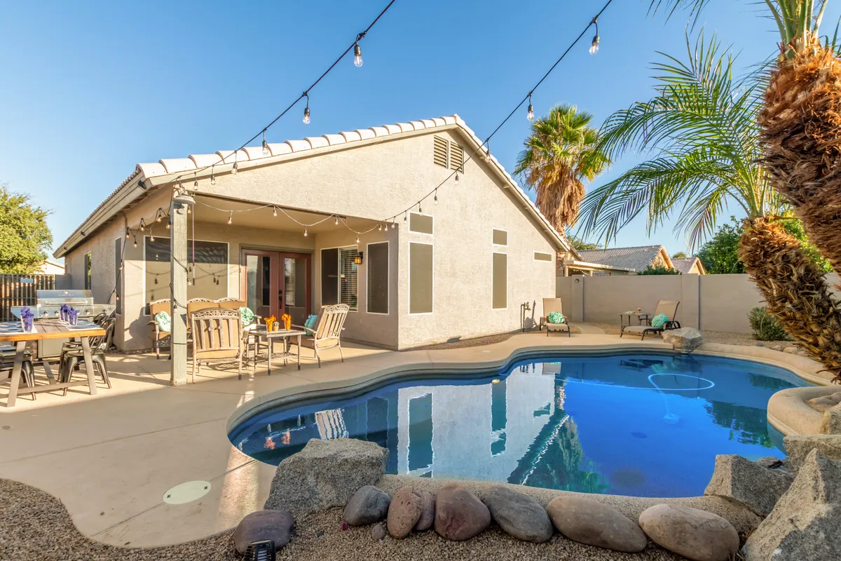 A wide-angle view of the swimming pool bordered by outdoor seating, palm trees, and atmospheric string lights. The scene is lively and cheerful, emphasizing space for swimming, barbecuing, and starlit outdoor dining. The layout shows ample room for both relaxation and social activities.