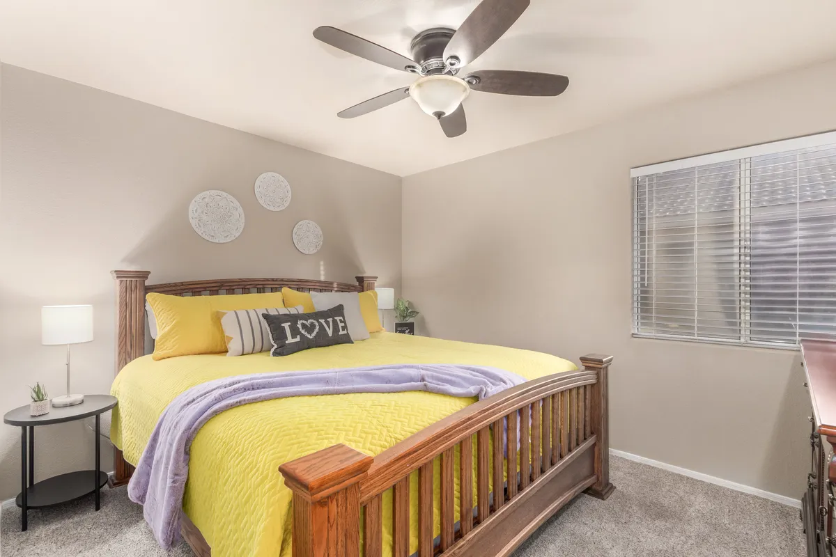A comfortable bedroom with a bed dressed in yellow and gray bedding, decorative pillows spelling 'LOVE,' a ceiling fan above, and a cozy nightstand with a lamp. The room exudes warmth and tranquility, ideal for winding down after a busy day.