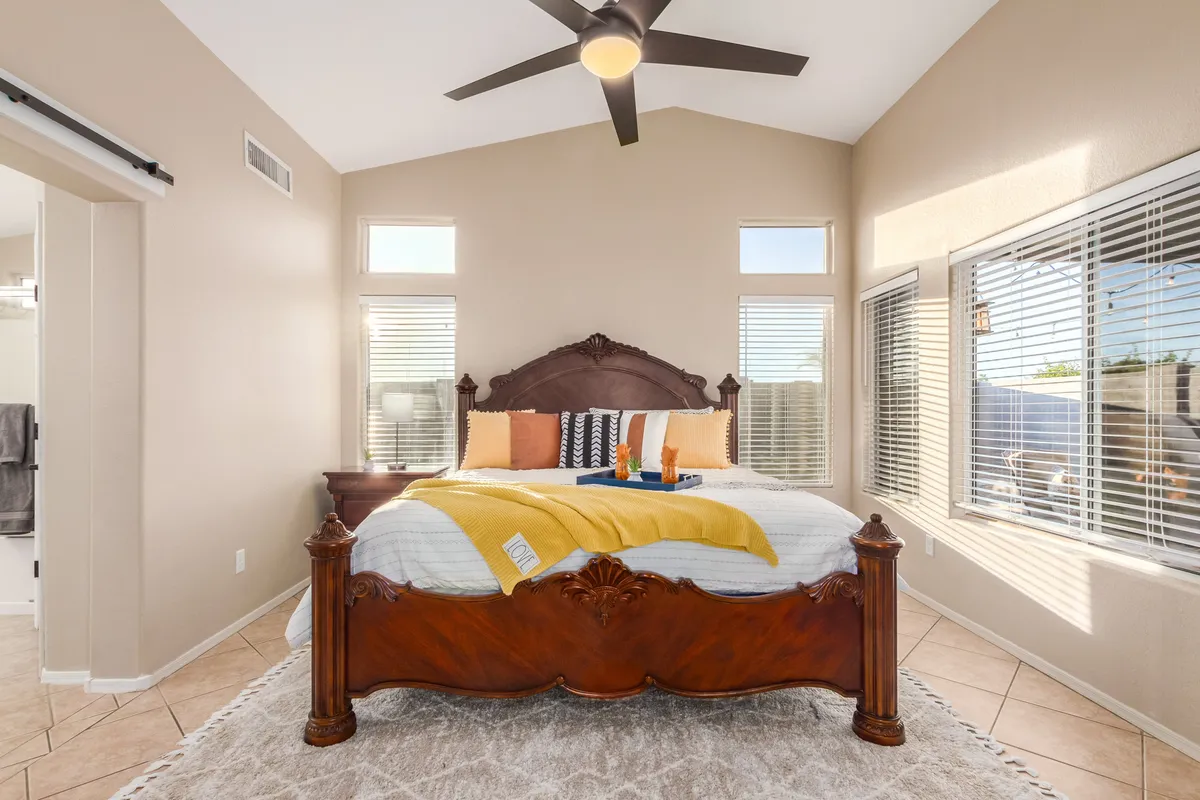 A bright, cozy bedroom featuring an ornate wooden bed frame adorned with colorful decorative pillows and a yellow blanket. Large windows flood the room with natural light, creating a warm, inviting ambiance that emphasizes relaxation and comfort.