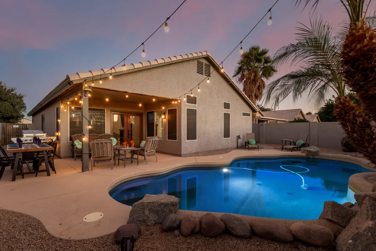 This image captures a sparkling swimming pool with a spacious patio seating area lit by string lights. The pool is surrounded by comfortable chairs and lush landscaping, creating a friendly and welcoming atmosphere for gatherings or quiet relaxation. The view highlights how the outdoor space can transition from sunny days to cozy evenings.